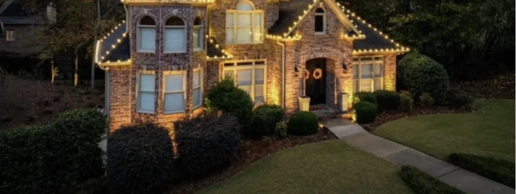 Brick home with gabled dormers outlined in warm white lights above a trimmed lawn at night.