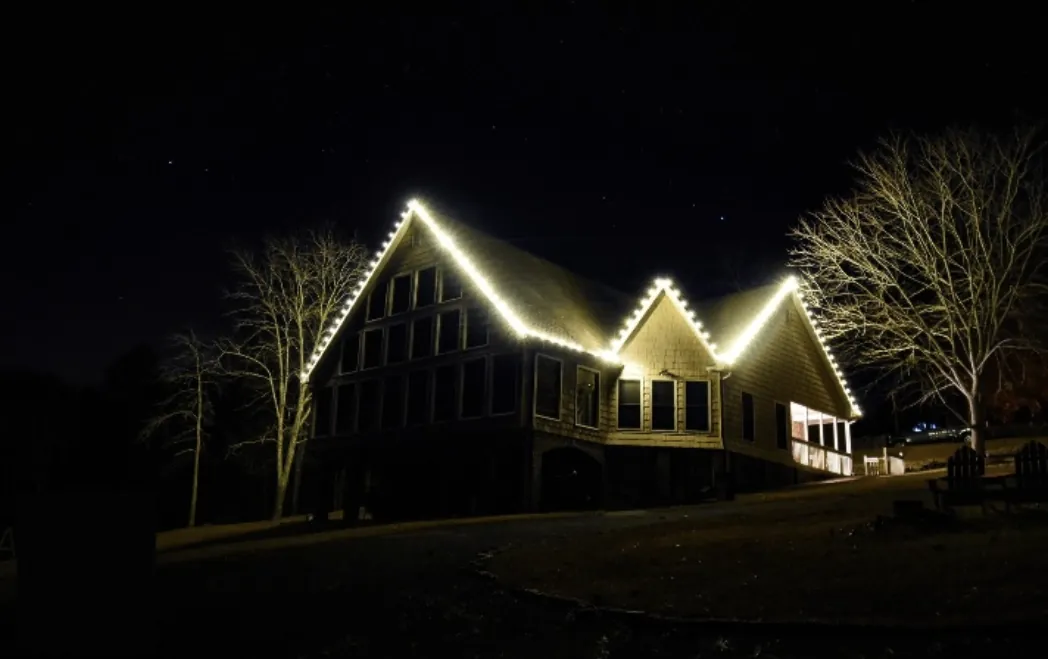 Wooded-lot home with three lit gables stacked along the roofline against a dark sky.
