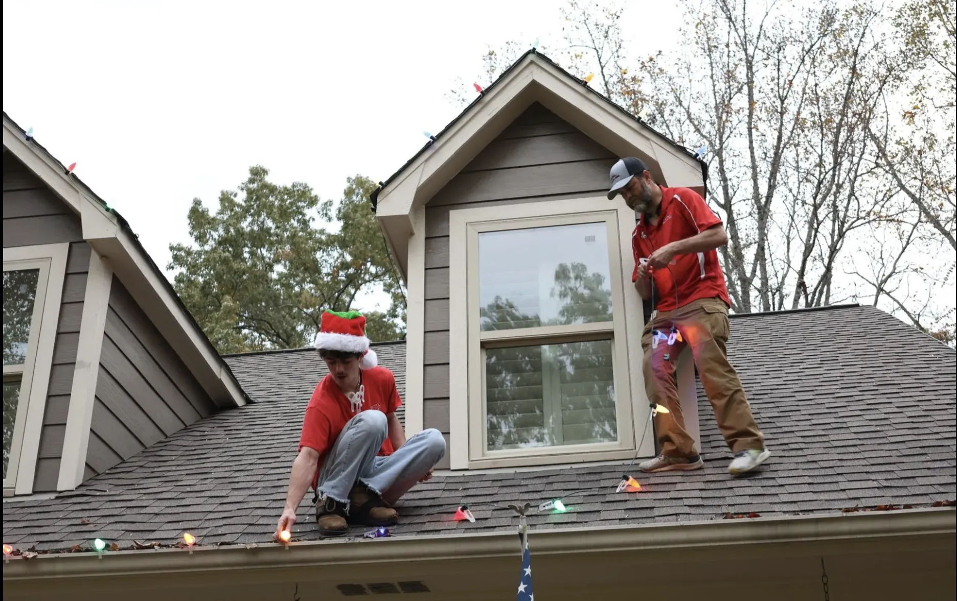 Two installers on a shingle roof beside a dormer, clipping a strand of lights along the eave.
